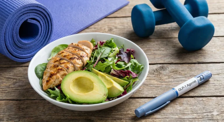 A bowl of grilled chicken salad with avocado on a wooden table, next to a rolled yoga mat, two blue dumbbells and an unbranded GLP-1 agonist injection pen.
